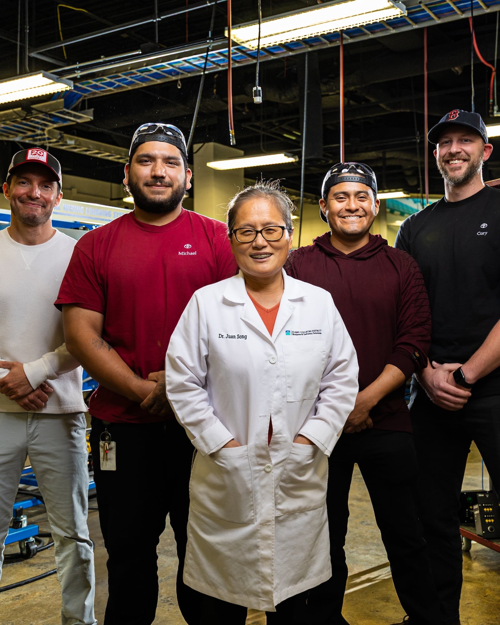 A diverse group of five people stands confidently in a workshop. They're wearing casual and lab attire, surrounded by industrial equipment, conveying teamwork.