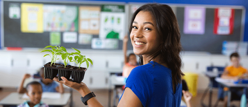 smiling teacher holding a plant and instructing elementary age students