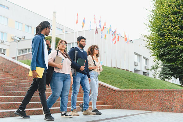a multicultural group of students walk across a campus talking to one another; several national flags can be seen flying on flag poles in the background