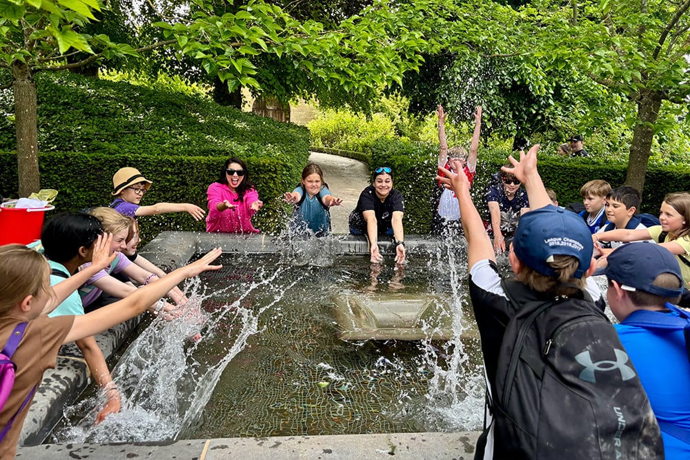 Children and adults laugh and splash around a bubbling outdoor fountain in a leafy garden, raising their hands in a joyful summer scene.