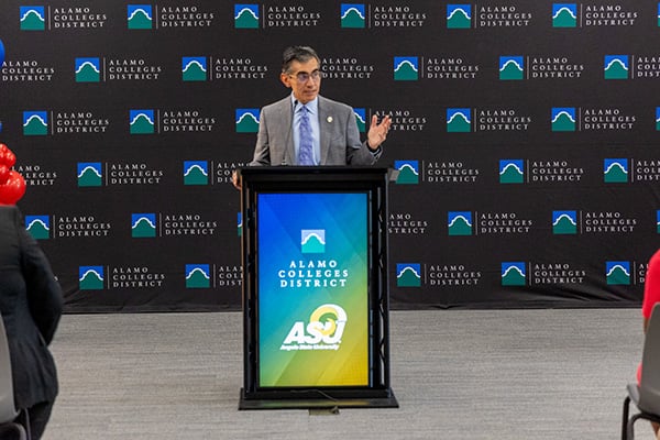 ACD Chancellor, Dr. Mike Flores, speaks at an Alamo Colleges District podium, with an ASO logo and branded backdrop behind him; audience members flank him.