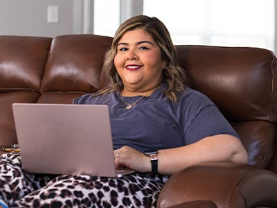 Kristen De Los Santos sitting on the couch with her laptop. Smiling at the camera.