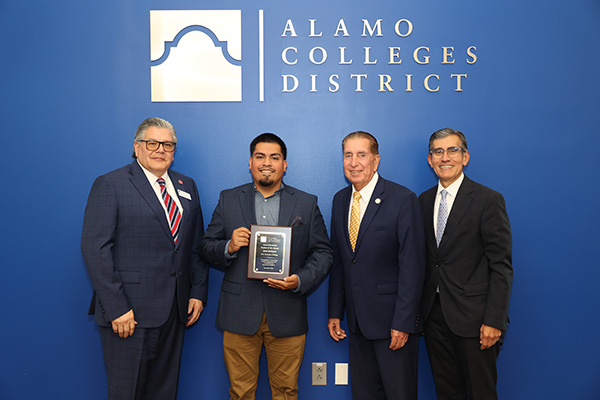 Jason Rodriguez with two Alamo Colleges board members and Chancellor Mike Flores in business attire pose before an Alamo Colleges District backdrop; the center man holds an award plaque, conveying pride and celebration.